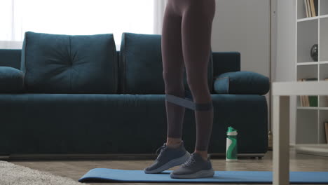 home-fitness-woman-is-training-legs-with-sporty-elastic-band-tensing-muscles-closeup-view-in-living-room-workout-and-exercise